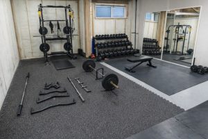 Hemmagym med skivstångsställning, hantlar och träningsbänk på gummimatta.